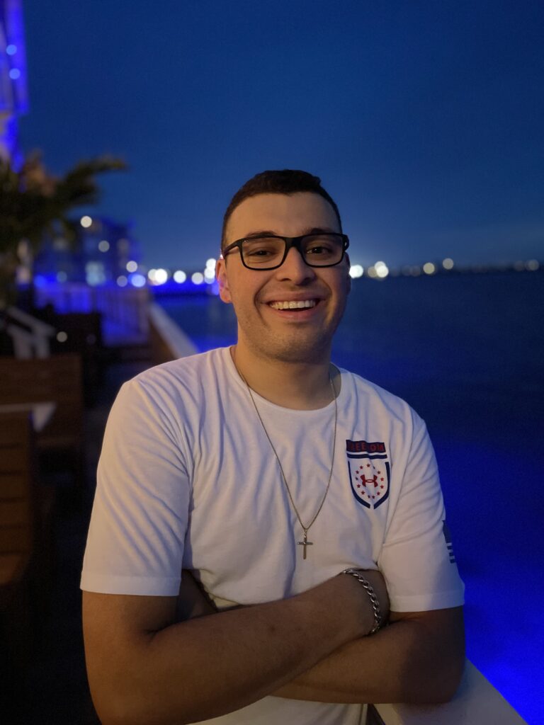 A picture of Bryan Rivas. He is Caucasian and has his arms crossed in front of him. He has short black hair and black glasses. His shirt is white with a logo on the top right and he has a cross necklace. A silver chain is on his right wrist. He appears to be by a body of water at night