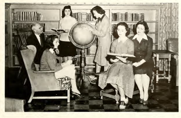 A black and white image of the International Relations Club. There are five women and the photo and one man. The man sits in the back left corner. To his right is a woman holding open a book and another looking at a globe. In the front two women look at the same book and to the left of them the final one sits perched in her chair.