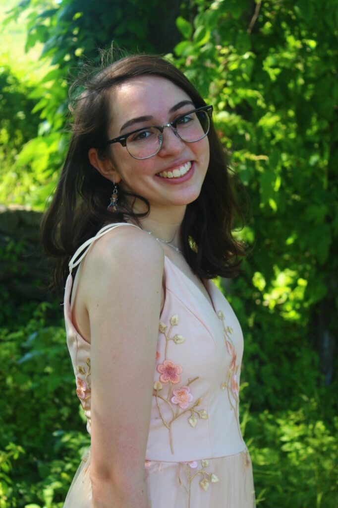 A picture of Eliza Vegas. She is Caucasian and wearing a white dress with pink flowers on it. She has in earrings and has black glasses. She is smiling at the camera.