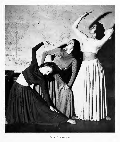 Three woman stand in a row in a black and white image. The first one is wearing a black shirt with long sleeves and a long black skirt. One leg is stretched out to the right and her arm is reaching over her head. Behind her stands a woman in gray with her hands on her knees. In the back is a woman in white. She has one arm extended slightly upward, bent towards the right. 